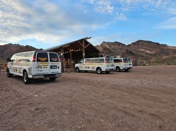 Three white tour vans parked on a gravel lot beside a rustic wooden barn with string lights, framed by rugged desert canyon cliffs under a wide blue sky.