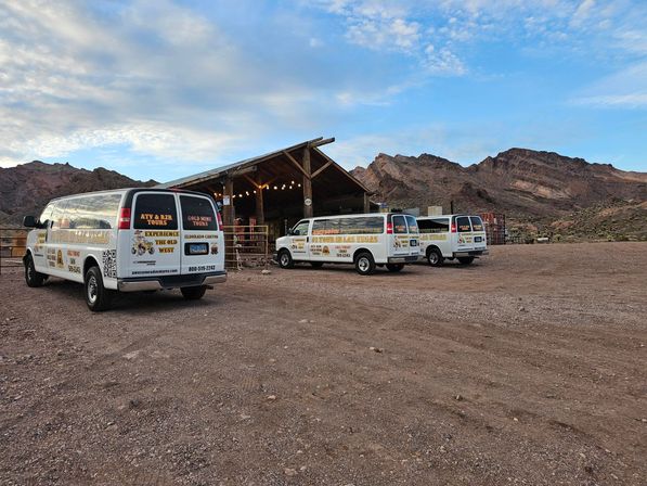 Three white tour vans parked on a gravel lot beside a rustic wooden barn with string lights, framed by rugged desert canyon cliffs under a wide blue sky.