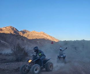 Two ATV riders kicking up dust on a desert off‑road trail with red safety flags, rugged sunlit mountains and a clear blue sky