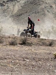 Off‑road ATV rider in a pink helmet and red gloves kicking up dust across rocky desert terrain with scrub brush and arid hills in the background