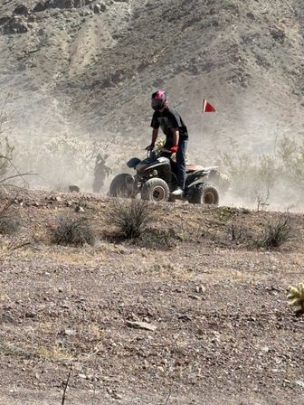 Off‑road ATV rider in a pink helmet and red gloves kicking up dust across rocky desert terrain with scrub brush and arid hills in the background