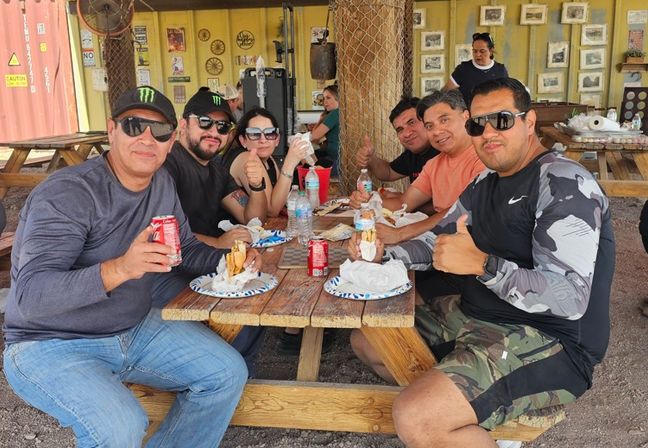 Six adults in sunglasses and caps share sandwiches and soda at a wooden picnic table in a shaded outdoor patio with rustic decor, smiling and giving thumbs-up.