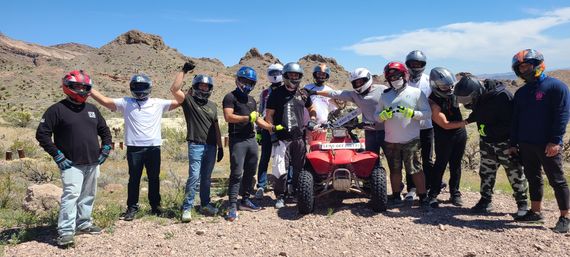 Group of helmeted riders posing with a red ATV on a sunny desert trail amid rocky hills and cacti.