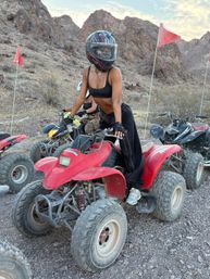 Rider in a full-face helmet and black top perched on a red ATV among rocky desert canyon terrain with other quad bikes, red safety flags, and a pastel evening sky.
