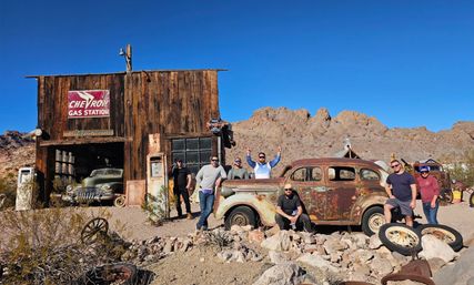 Group of visitors posing by a rusted vintage car and old pumps in front of a weathered wooden desert gas station amid rocky mountains under a clear blue sky