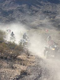 Group of ATV riders on quad bikes tearing along a dusty desert trail with red safety flags and rugged scrubby hills in the background.