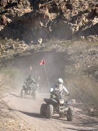 Helmeted riders on ATVs kicking up dust on a rocky desert canyon trail with orange safety flags — off‑road quad biking adventure.