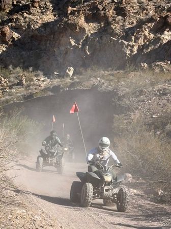 Helmeted riders on ATVs kicking up dust on a rocky desert canyon trail with orange safety flags — off‑road quad biking adventure.