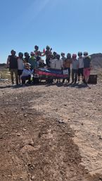 Group of off-road riders in helmets posing with a banner on a sunny, rocky desert trail with distant hills