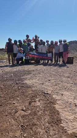 Group of off-road riders in helmets posing with a banner on a sunny, rocky desert trail with distant hills