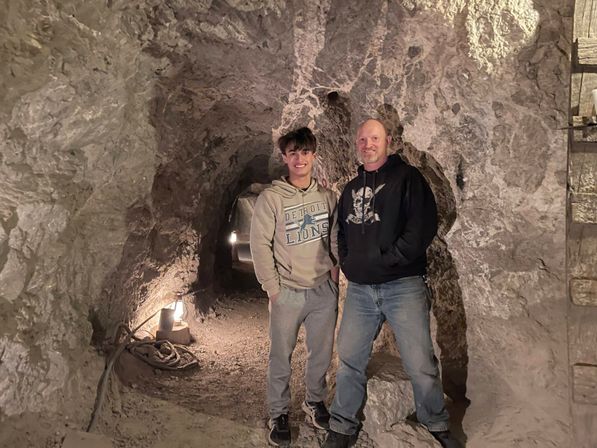 Two visitors in hoodies smiling inside a dimly lit underground mine tunnel with rugged stone walls, gravel floor, rope and lantern creating a cozy cave atmosphere.