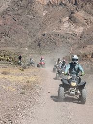 Group of helmeted ATV riders kicking up dust on a rocky desert trail below rugged mountains — off-road quad adventure in arid canyon terrain