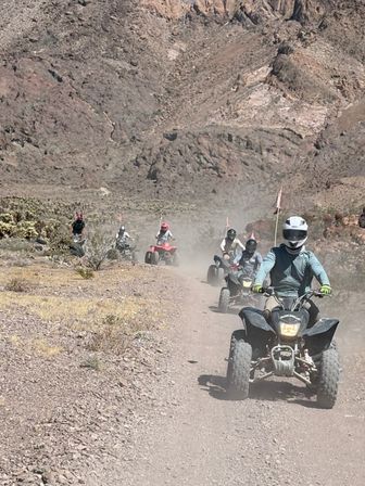 Group of helmeted ATV riders kicking up dust on a rocky desert trail below rugged mountains — off-road quad adventure in arid canyon terrain