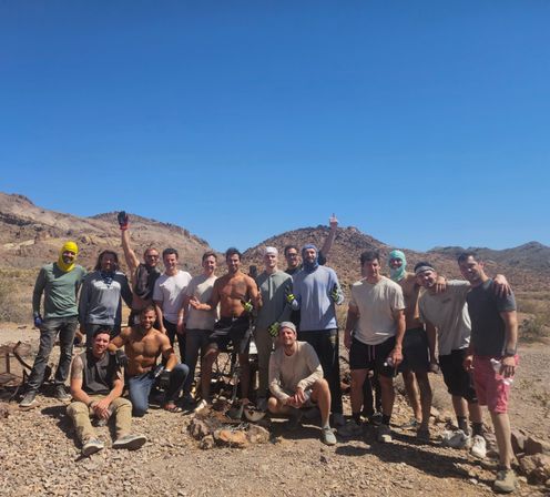 About 15 men posing together in a rocky desert landscape with low mountains and a clear blue sky, wearing casual outdoor gear, bandanas and gloves, some shirtless and smiling after a day of outdoor work.