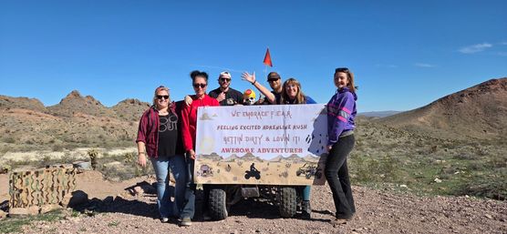 Group of seven people posing with an ATV in a rocky desert holding a banner that reads We Embrace F.E.A.R. — Feeling Excited Adrenaline Rush, Gettin Dirty & Lovin It! Awesome Adventures, clear blue sky and rugged hills in background