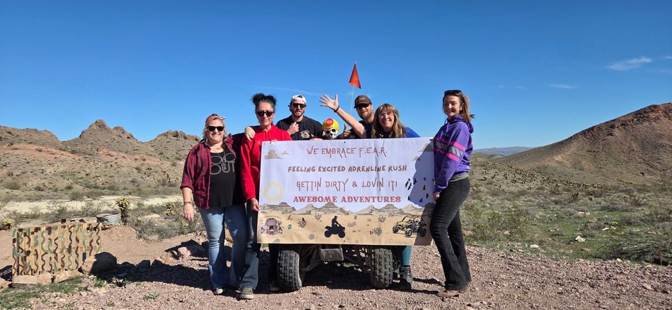 Group of seven people posing with an ATV in a rocky desert holding a banner that reads We Embrace F.E.A.R. — Feeling Excited Adrenaline Rush, Gettin Dirty & Lovin It! Awesome Adventures, clear blue sky and rugged hills in background