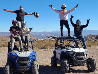 Four people in helmets with arms raised posing on two side-by-side off-road UTVs in a desert mountain landscape under a clear blue sky