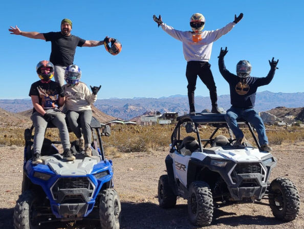 Four people in helmets with arms raised posing on two side-by-side off-road UTVs in a desert mountain landscape under a clear blue sky
