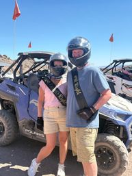 Couple in helmets wearing playful 'Mr Right' and 'Mrs Always Right' sashes pose next to off‑road UTVs on a sunny desert trail with orange safety flags.
