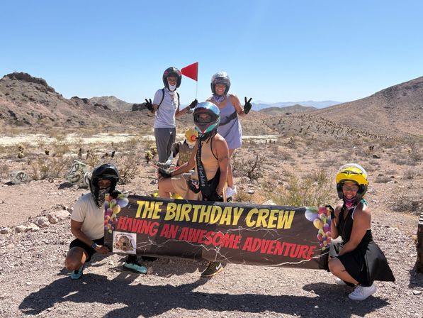 Five people in helmets and off-road gear posing with a colorful banner reading "THE BIRTHDAY CREW – HAVING AN AWESOME ADVENTURE" in a sunny desert landscape with rocky hills and clear blue sky.