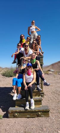 Cheerful group of friends wearing bandanas and sunglasses stacked on a metal structure in a sunny desert setting with clear blue sky — outdoor hiking group photo.