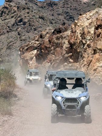Three side-by-side off-road UTVs kicking up dust on a narrow desert canyon trail beneath rugged rocky cliffs, drivers wearing helmets and safety flags visible