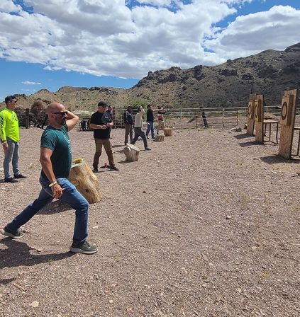 Group of people axe throwing at an outdoor range in a rocky desert valley, wooden targets lined up against a mountain backdrop under a blue sky with scattered clouds.