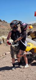 Desert ATV elopement: two helmeted riders embrace, tattooed woman in a veil holding a bouquet beside a yellow quad and a skeleton mannequin in a hard hat against a rocky desert backdrop.