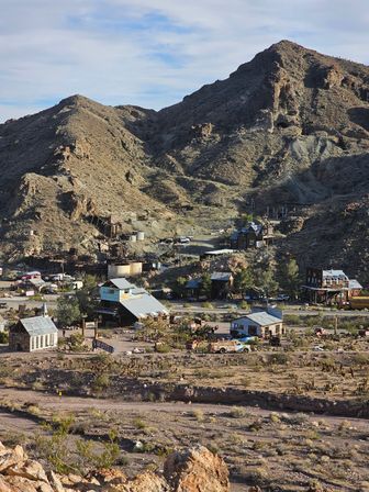 Sunlit desert ghost town in an arid mountain valley with old mining ruins, weathered wooden buildings, rusted vehicles and a tilted motel sign