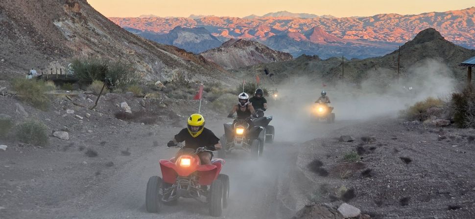 Group of ATV riders on a dusty desert trail through a rocky canyon at sunset with pink-lit mountains in the background