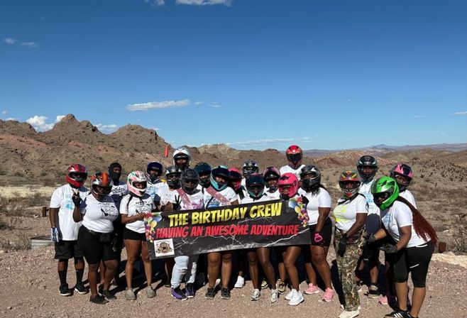 Group of helmeted off-road riders holding a banner reading “The Birthday Crew” on a sunny desert trail with rocky hills and clear blue sky