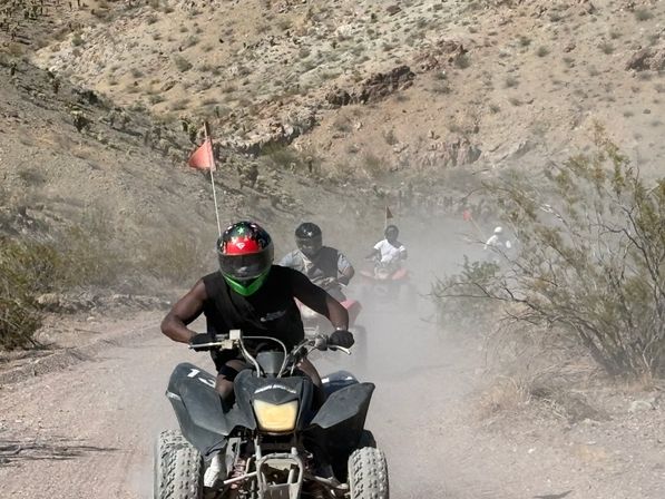 Three helmeted ATV riders tearing down a dusty rocky desert canyon trail, kicking up dirt past scrub brush and orange safety flags