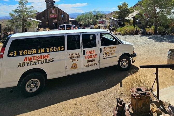 White Las Vegas tour van parked in a rustic desert ghost town with old wooden mine buildings, scattered trees, dusty courtyard, and mountains in the background.