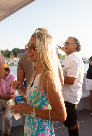 Smiling woman in a colorful summer dress and sunglasses holding a water bottle and phone aboard a sunny harbor boat cruise with marina and passengers in the background