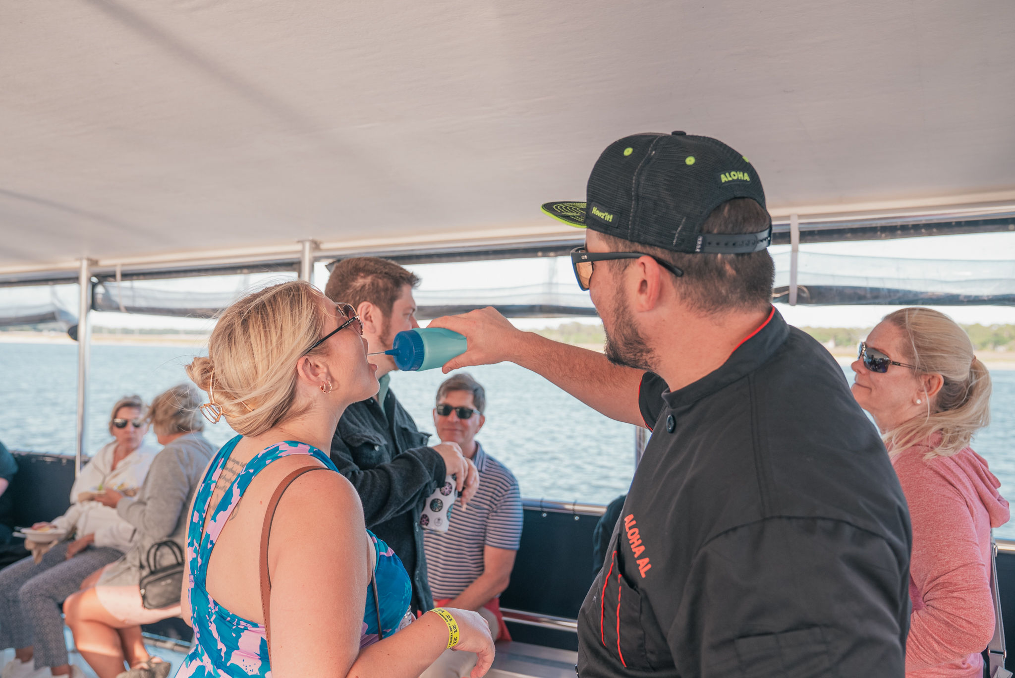 Man playfully pours a drink from a blue shaker into a woman's mouth aboard a daytime boat cruise, with seated passengers, calm water and a distant shoreline visible.