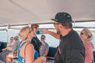 Man playfully pours a drink from a blue shaker into a woman's mouth aboard a daytime boat cruise, with seated passengers, calm water and a distant shoreline visible.