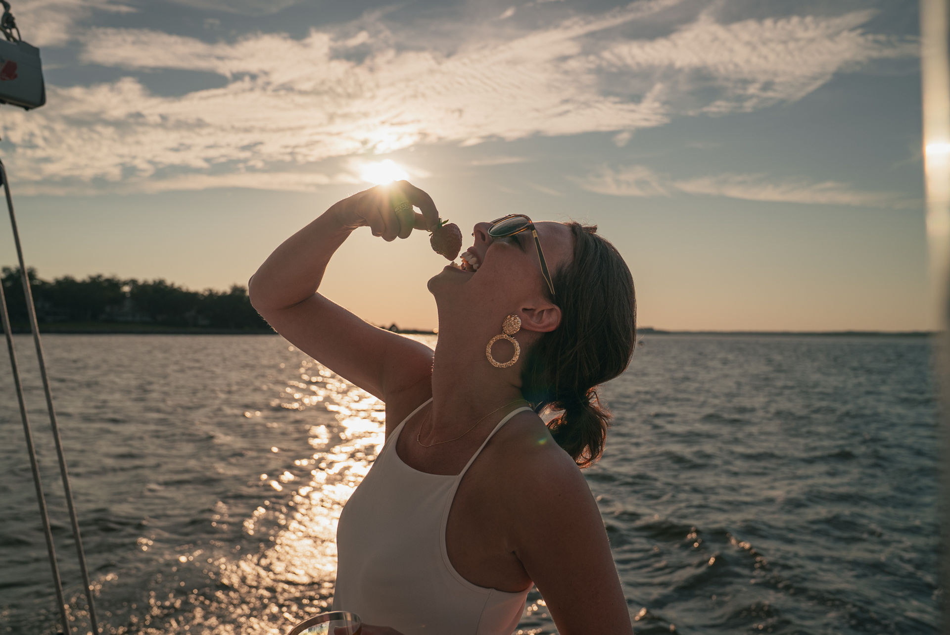 Woman on a sailboat at sunset wearing sunglasses and gold hoop earrings, biting a chocolate-covered strawberry with sunlight sparkling on coastal water