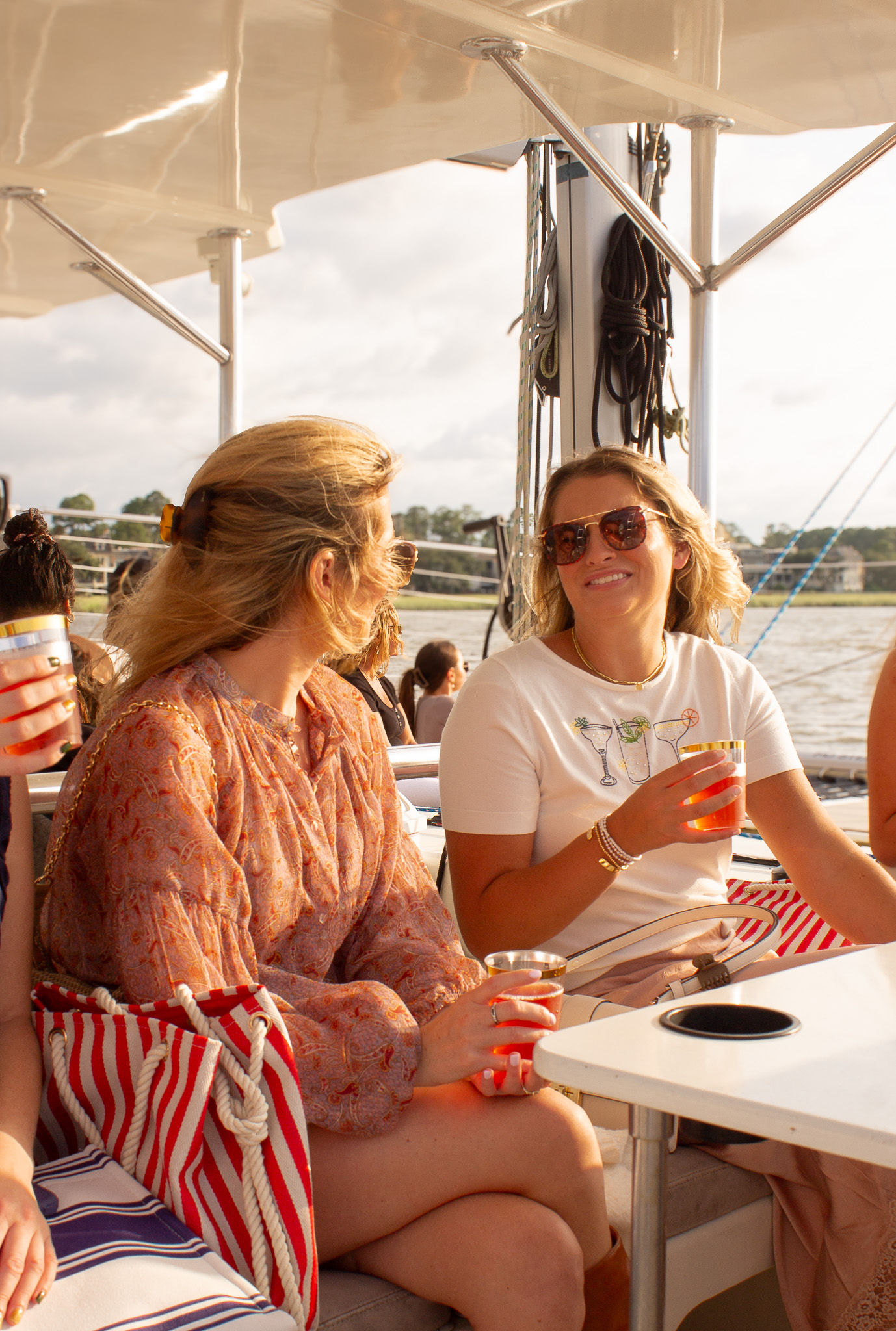 Two women chatting and smiling on a sunlit sailboat deck during a summer cruise, holding cold drinks with a red-and-white striped beach bag and marina waterfront in the background.