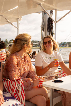 Two women chatting and smiling on a sunlit sailboat deck during a summer cruise, holding cold drinks with a red-and-white striped beach bag and marina waterfront in the background.