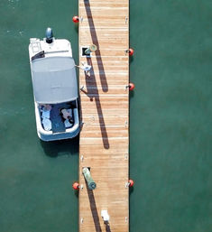 Aerial view of a motorboat moored alongside a wooden dock with orange buoys and two people relaxing on board over green water