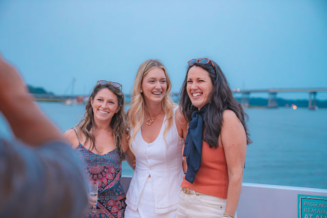 Three laughing women in summer outfits posing on a boat deck at a coastal harbor at dusk, with a bridge and sailboats on calm water in the background