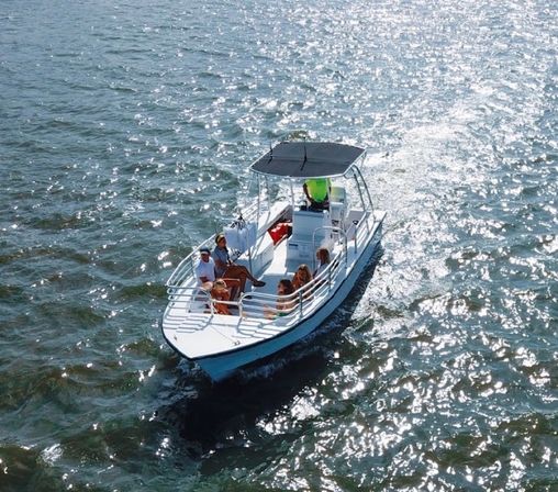 Aerial view of a small white motorboat carrying a group of people enjoying a sunny cruise across sparkling coastal waters under a shaded canopy.
