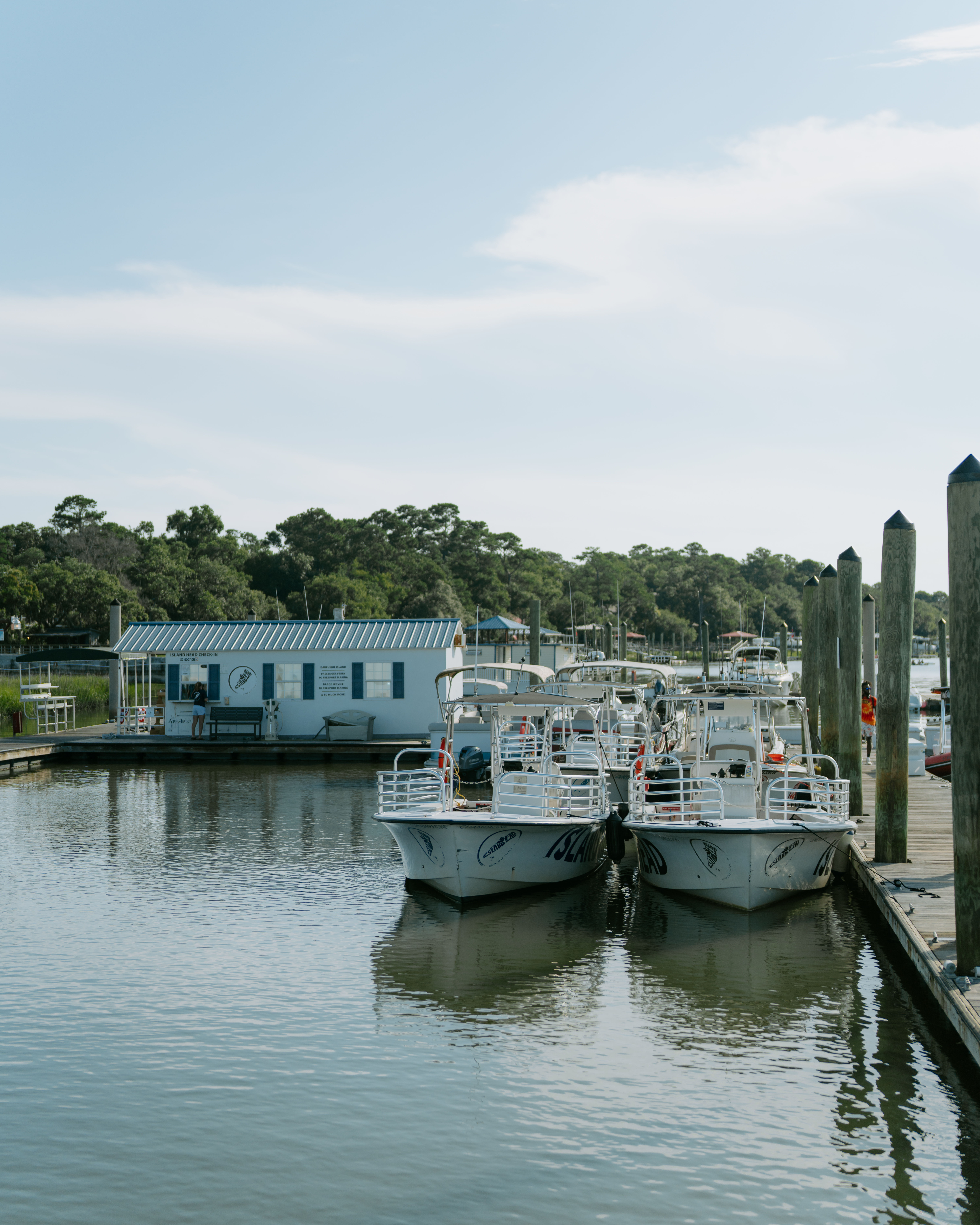 Two white boats moored side-by-side at a wooden dock in a peaceful coastal marina, calm reflective water, tree-lined shoreline and clear blue sky.