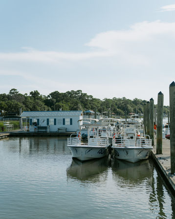 Two white boats moored side-by-side at a wooden dock in a peaceful coastal marina, calm reflective water, tree-lined shoreline and clear blue sky.