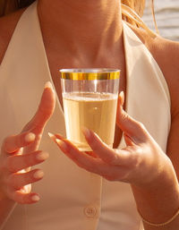 Sunlit close-up of a woman’s hands holding a clear cup with a gold rim filled with sparkling wine, cream halter top and beachside sunset glow