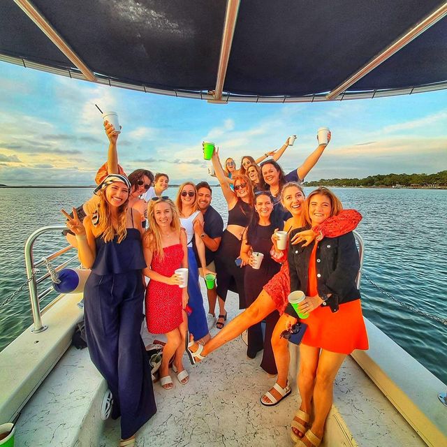 Boat party sunset cruise — cheerful group of friends on a boat deck raising cups and smiling over calm coastal waters.