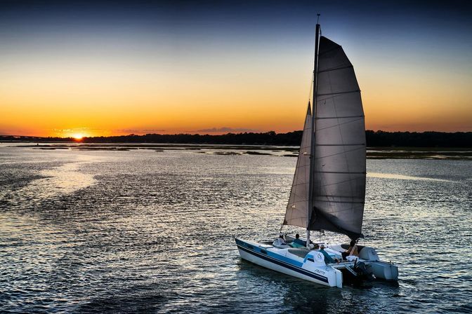 Catamaran sailboat gliding across a calm coastal estuary at sunset, orange-to-blue sky reflecting on rippled water with marshy shoreline on the horizon.