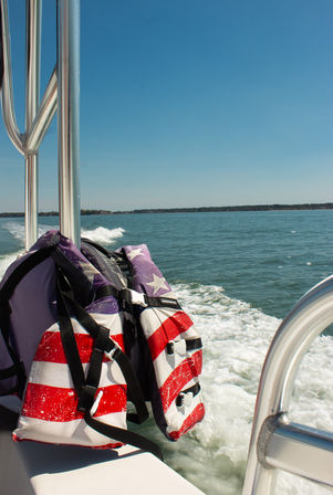 American flag life jacket draped on a motorboat rail as the boat leaves a white wake across a blue lake under a clear sunny sky