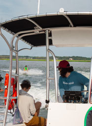 View from a covered motorboat towing a green-vested wakeboarder across a coastal inlet, with two people on deck, a life ring, boat wake, and marshy shoreline with houses in the distance.