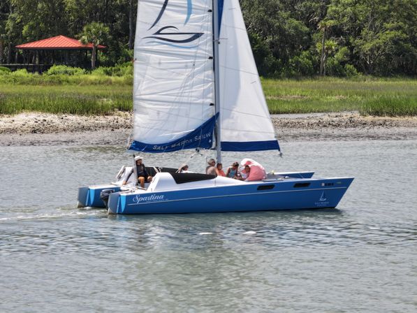 Blue catamaran sailboat with white sails and passengers cruising past a grassy marsh shoreline and a red-roof pavilion on a sunny day